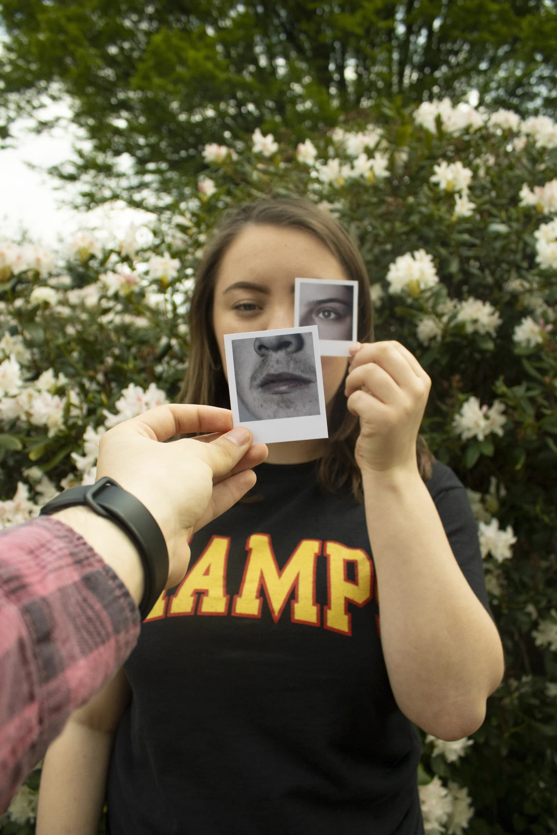 A person holds up two small Polaroid photographs in front of their face, aligning them with their own features. One photo shows their mouth and chin, held over their actual mouth and chin. The other photo shows their eye, positioned over their right eye.