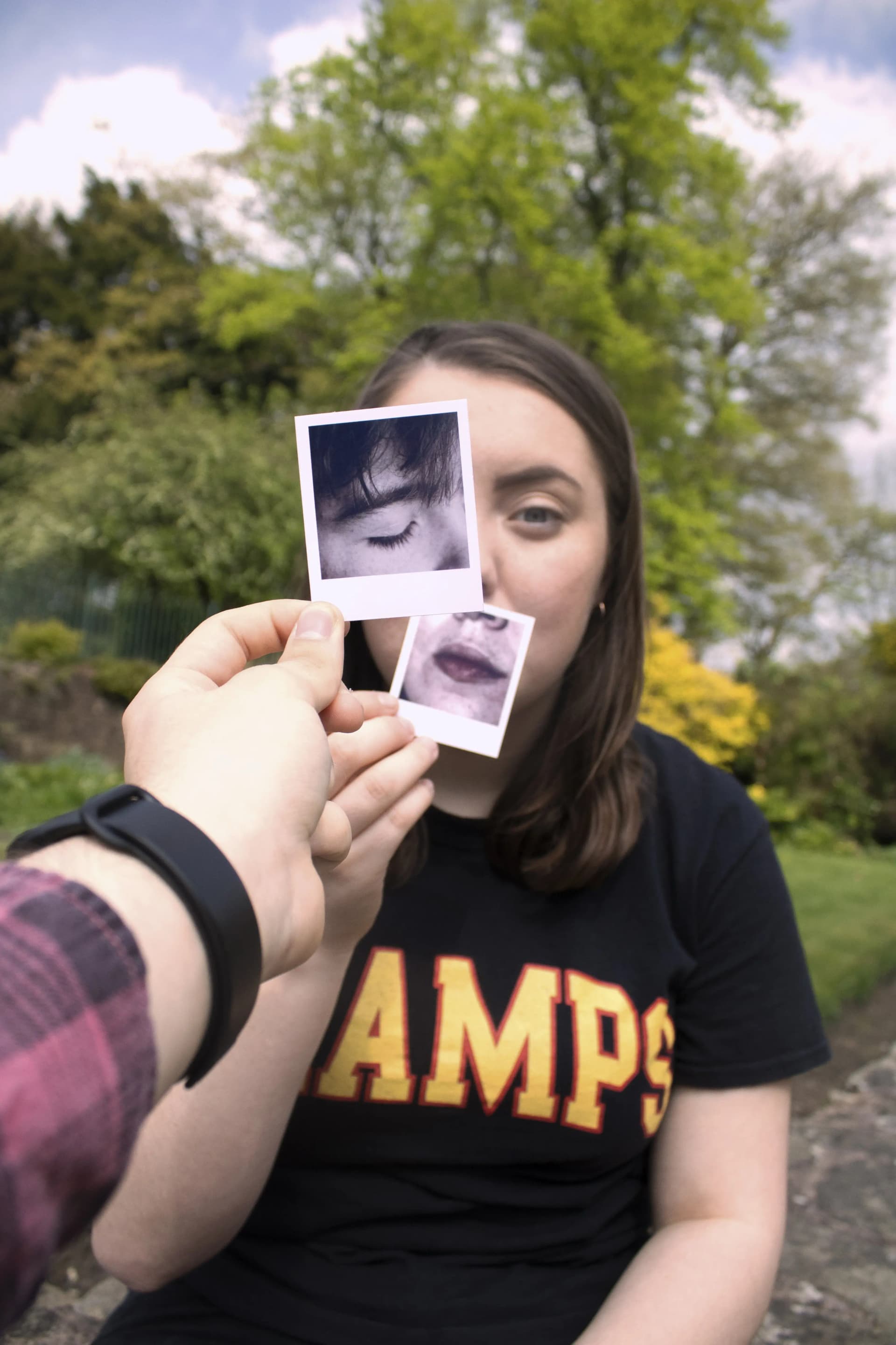 A person holds up two small Polaroid photographs in front of their face, aligning them with their own features. One photo shows their mouth and chin, held over their actual mouth and chin. The other photo shows their eye, positioned over their left eye.