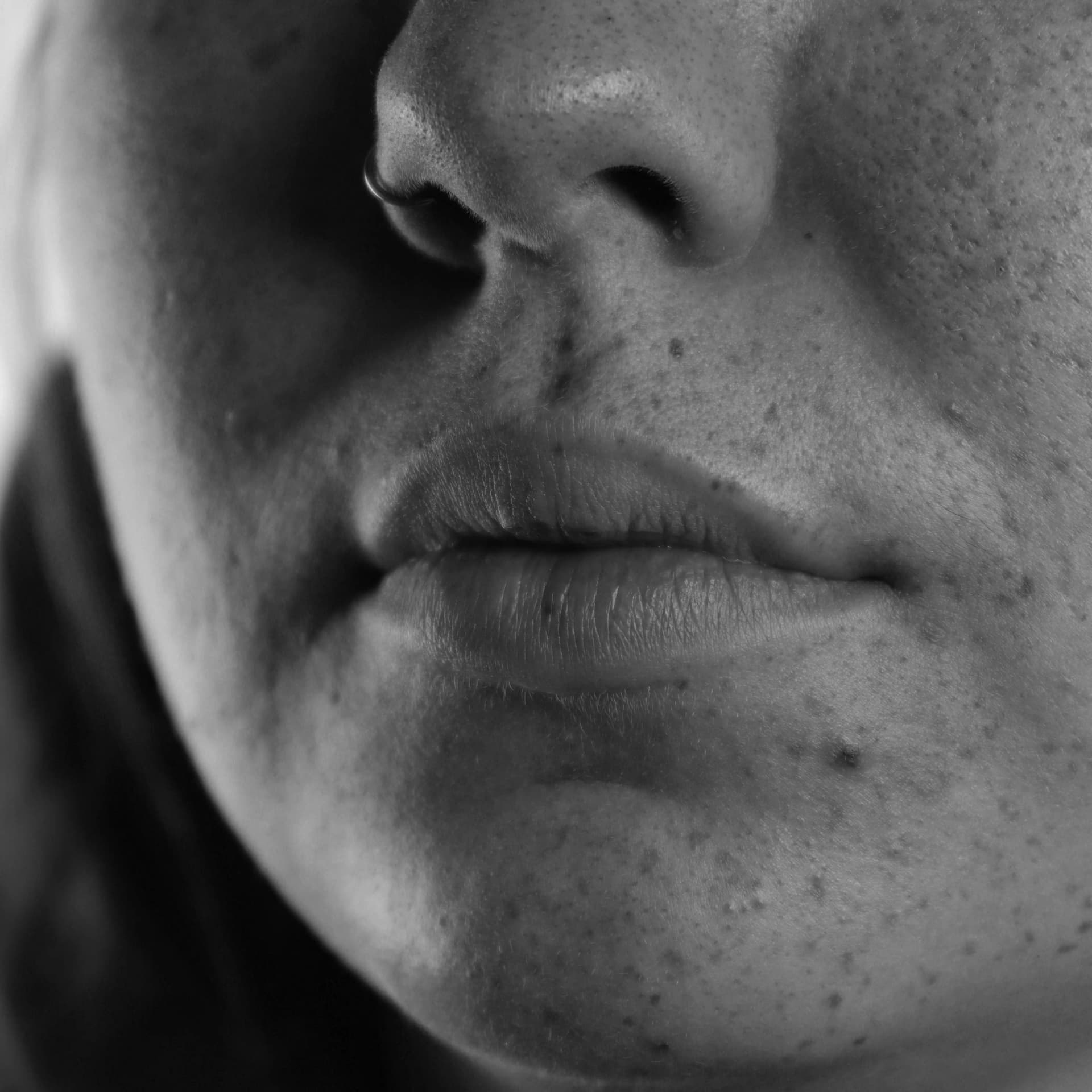 A detailed black and white close-up photograph focuses on the lower portion of a human face. The subject's full lips are centrally positioned, showing their texture and subtle contours. Above the lips, the base of a nose is visible, featuring a ring piercing in the left nostril. The surrounding skin exhibits a slightly uneven texture and a few small blemishes. The overall lighting highlights the subtle variations in tone and surface detail.